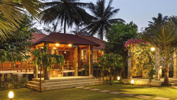 A well-lit, two-story house with a red roof, surrounded by lush greenery and palm trees. - Ananta Vilas by Stayjade (Bengaluru)