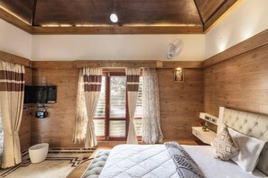 A bedroom with a wooden ceiling, a bed with a white quilt, a bedside table with a lamp, and a window with curtains. - Ananta Vilas by Stayjade (Bengaluru)