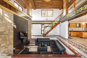A spacious living area with a stone wall, a large flat-screen TV, a black leather sofa, and a staircase leading to an upper level.