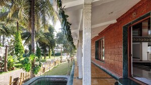 A corridor with a brick wall, a window, and a view of a garden with palm trees and a water feature. - Ananta Vilas by Stayjade (Bengaluru)