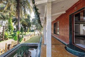 A corridor with a brick wall, a window, and a view of a garden with palm trees and a water feature. - Ananta Vilas by Stayjade (Bengaluru)