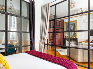 A hotel room with a large bed, a view of a red wall and a staircase, and a dining area visible through a glass partition. - Merveil Luxury Suites AboukirI-II-III-IV (Paris)