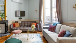 Un salon avec un canapé gris, un pouf rose et un tapis bleu. Un téléviseur est fixé au mur et il y a une fenêtre avec des rideaux.