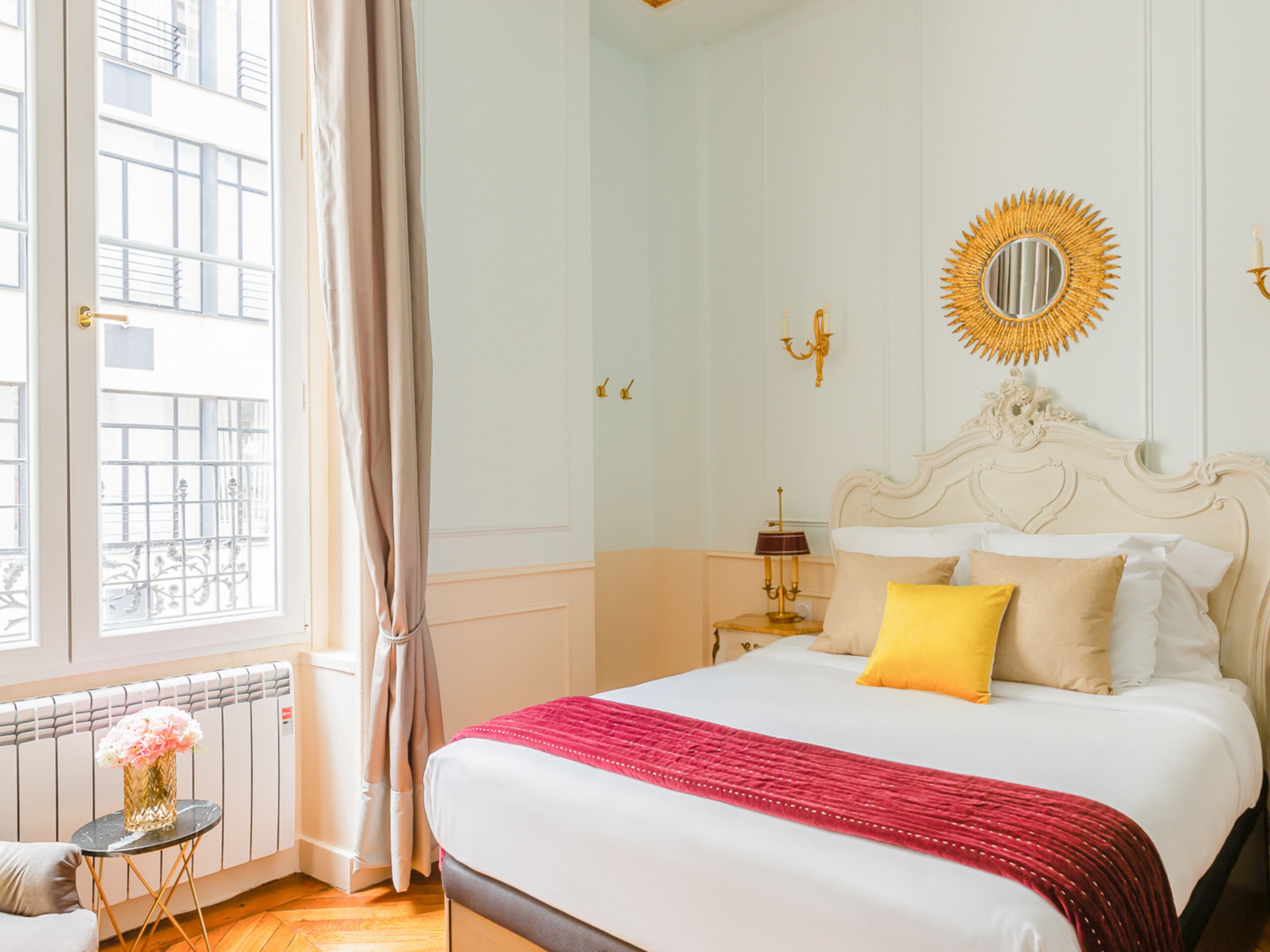 Ein Hotelzimmer mit einem großen Bett, einem runden Spiegel, einem kleinen Tisch mit einer Vase und Blick durch ein mit Vorhängen drapiertes Fenster hinaus.