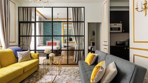Une chambre d’hôtel moderne avec un canapé jaune, un canapé gris et une grande fenêtre avec des rideaux. On y trouve un lit avec une tête de lit blanche et un mur bleu à motifs derrière. Une petite table avec un vase de fleurs est placée au centre de la pièce.