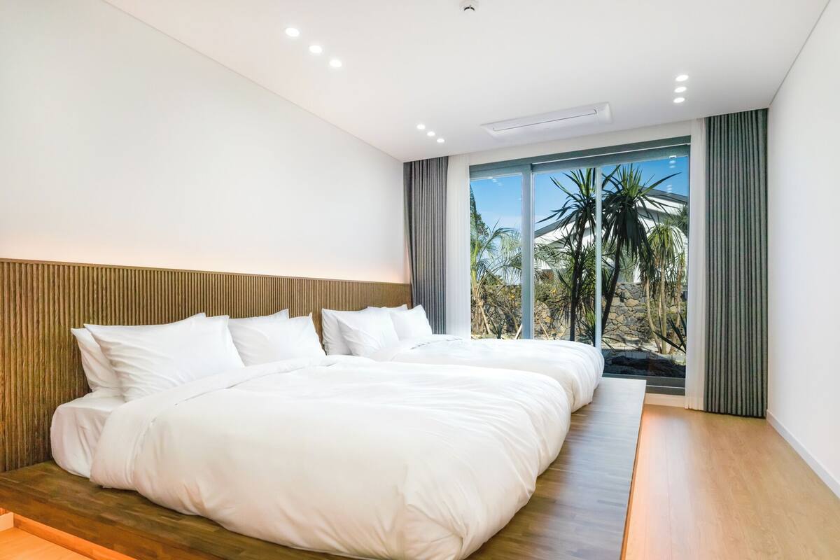 Habitación de hotel moderna con una cama grande, cabecera de madera y vista a palmeras y un muro de piedra.