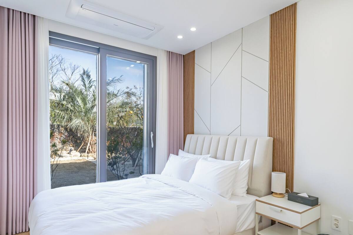 Un dormitorio moderno con una cama grande, una mesita de noche, una lámpara y vistas a palmeras a través de la puerta de vidrio.