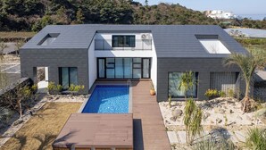 A modern two-story house with a swimming pool, a wooden deck, and a view of a hilly landscape. - Gobajinin Private Pool Villa (Jeju City)