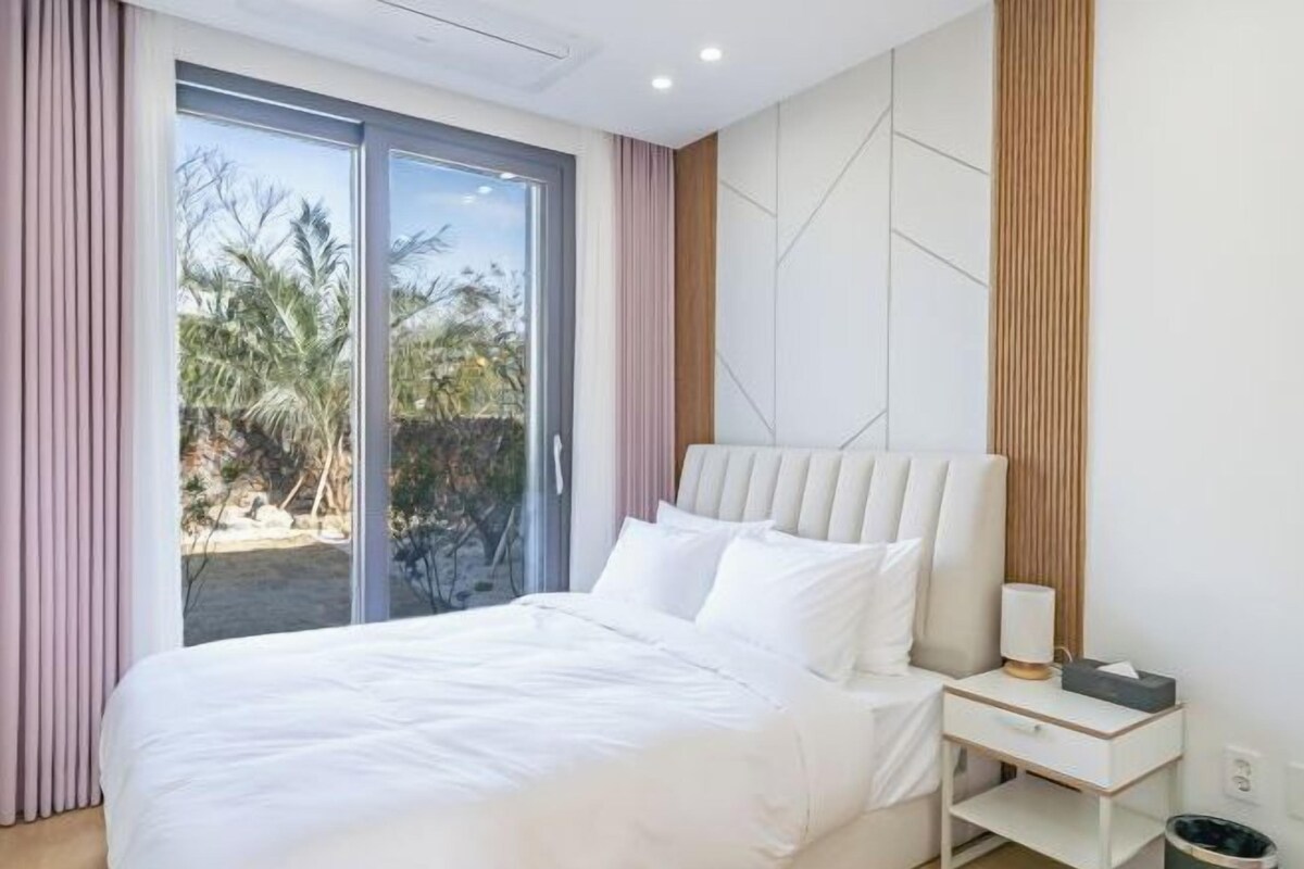 Une chambre moderne avec un grand lit, une table de chevet et une vue sur les palmiers, visible à travers une porte-fenêtre coulissante.