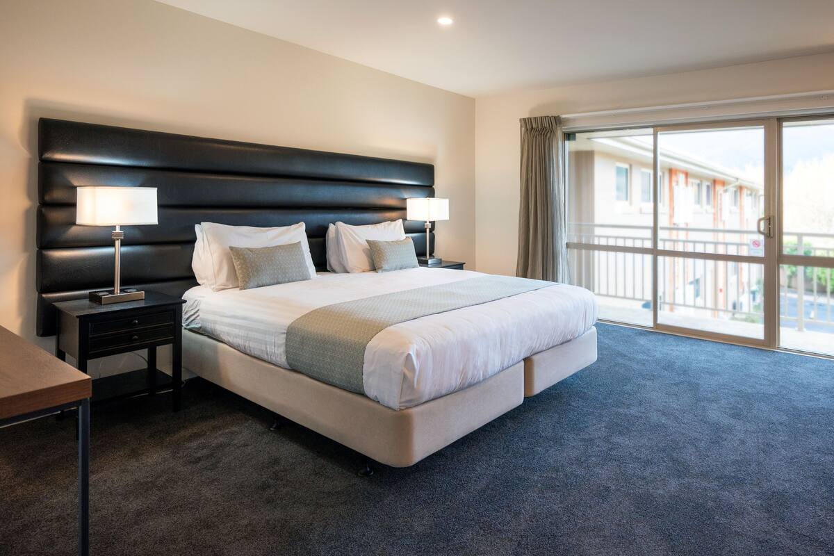 Habitación de hotel con una cama grande, dos mesitas de noche con lámparas, suelo alfombrado y un balcón con vistas.