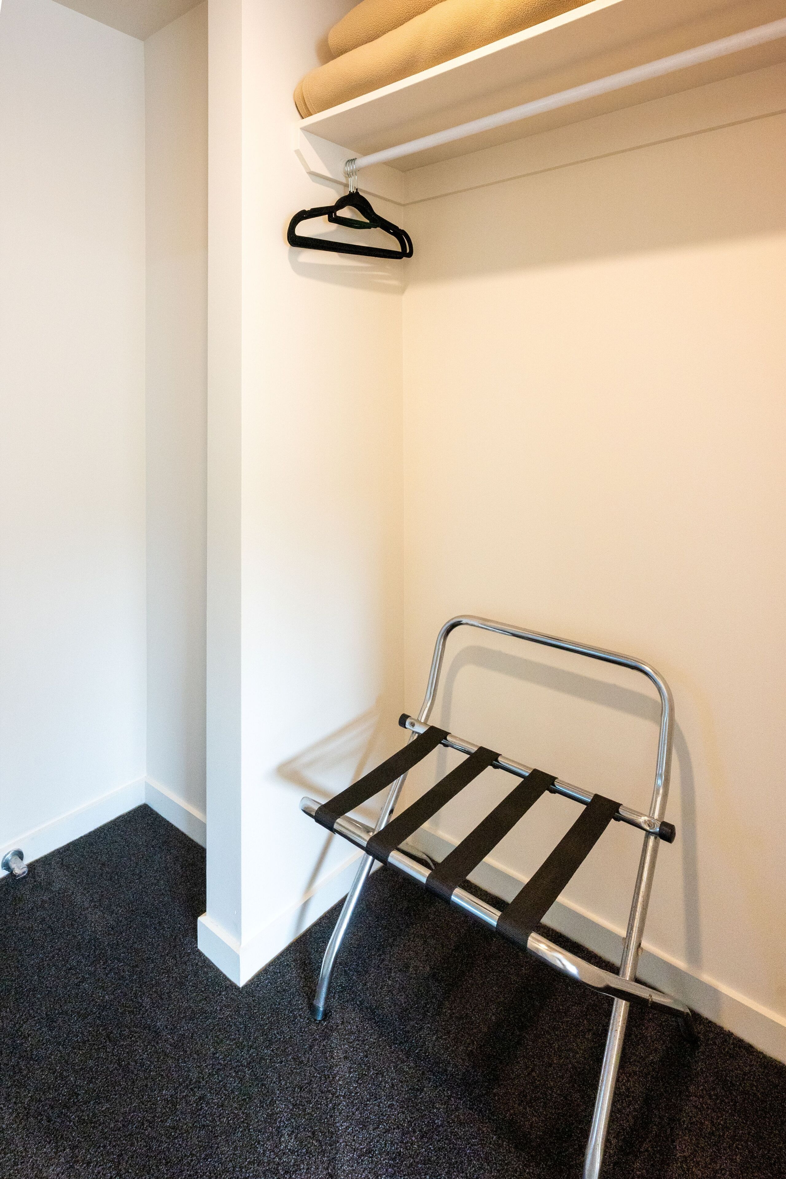 Un coin de pièce avec un étendoir métallique adossé à un mur blanc et un sol recouvert d’une moquette sombre.