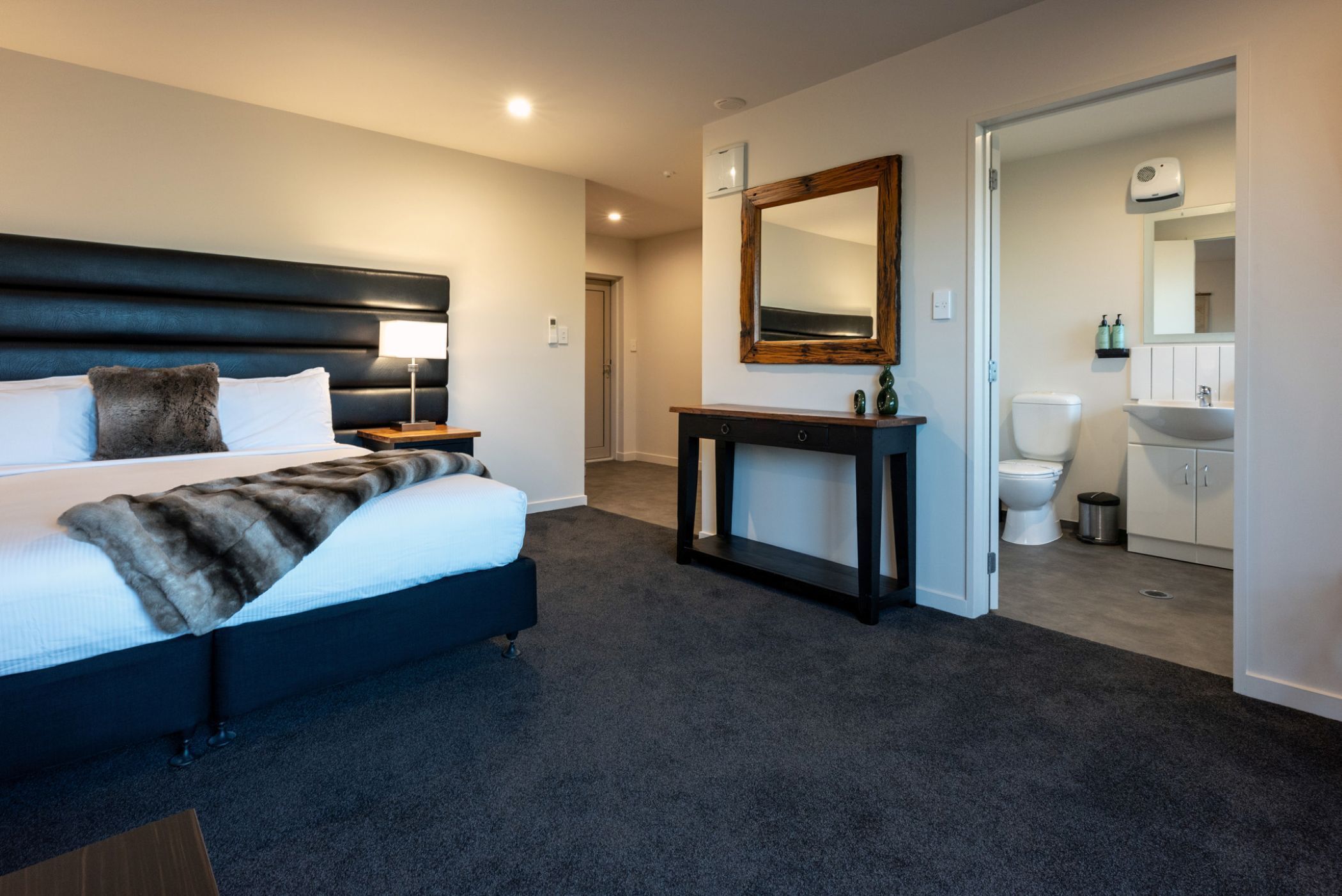 A modern hotel room with a large bed, a bedside table, a console table, a mirror, and a bathroom with a toilet and sink.