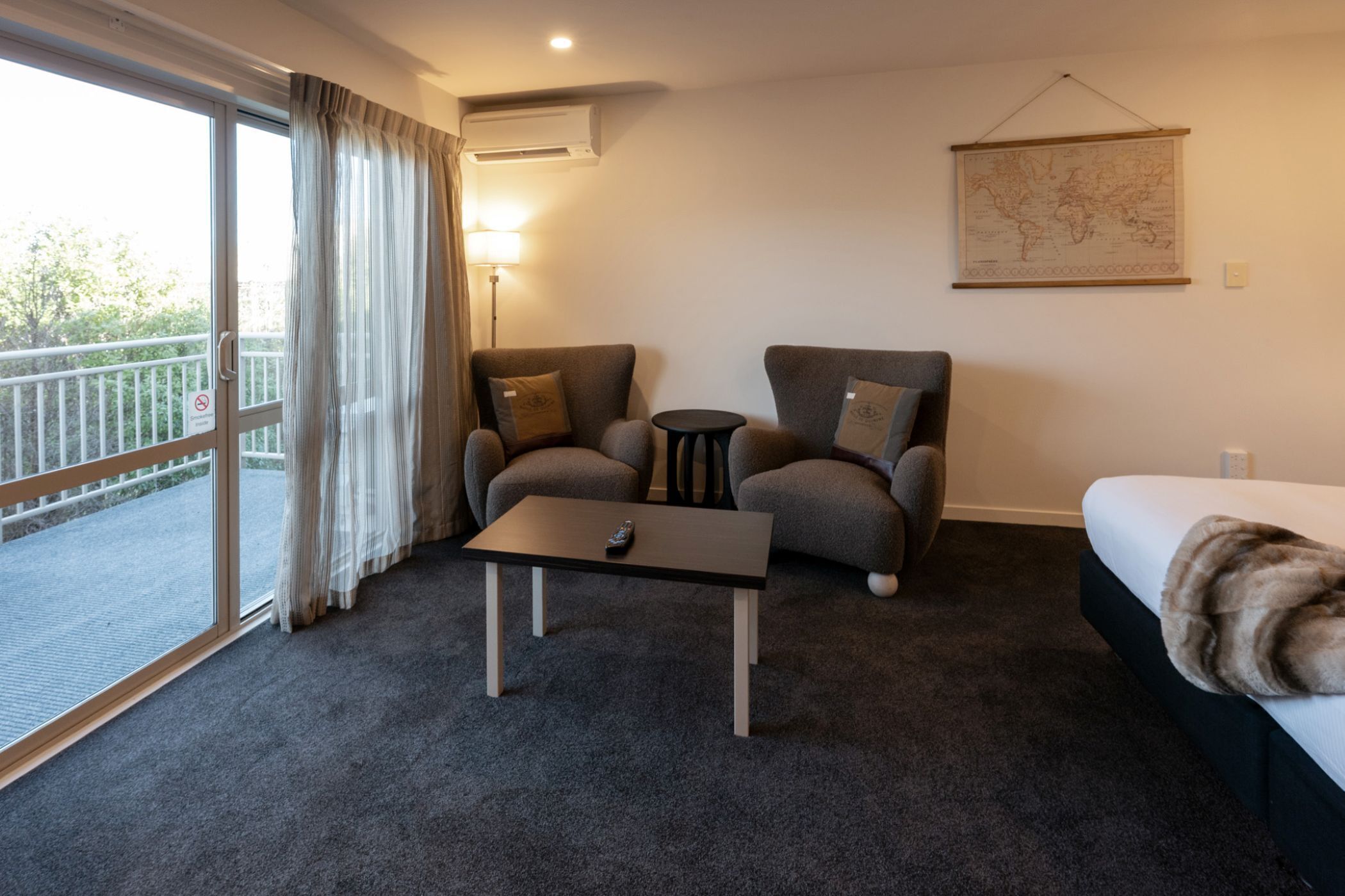 A hotel room with a bed, two armchairs, a small table, a map on the wall, and a balcony with a view.