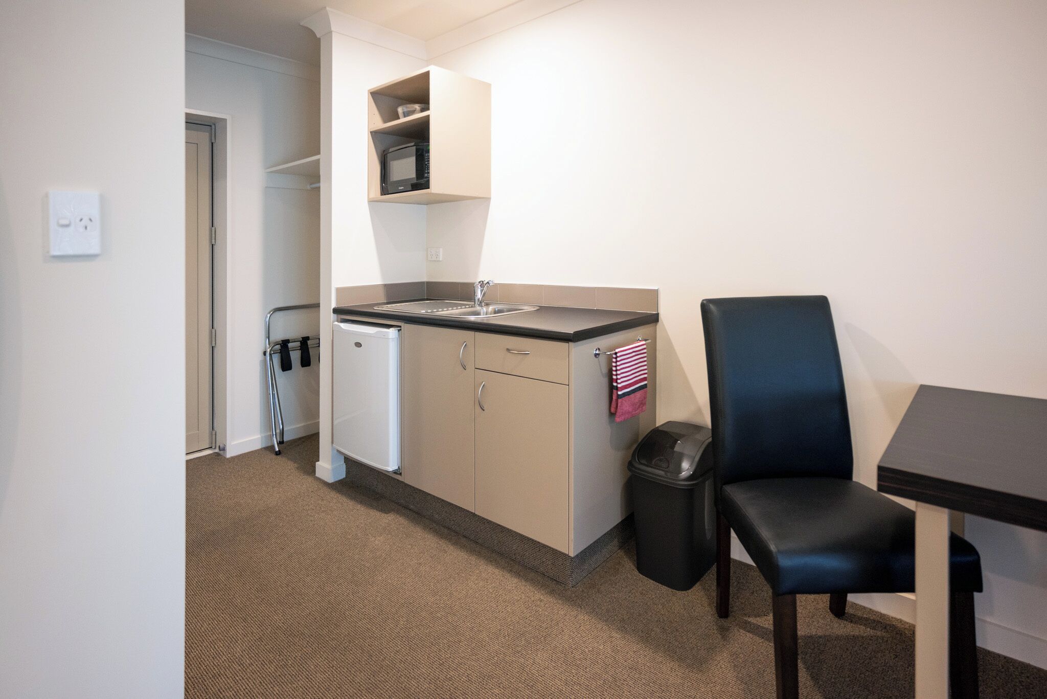 A compact kitchenette with a microwave, sink, and small refrigerator. There is a trash bin and a chair near the sink.