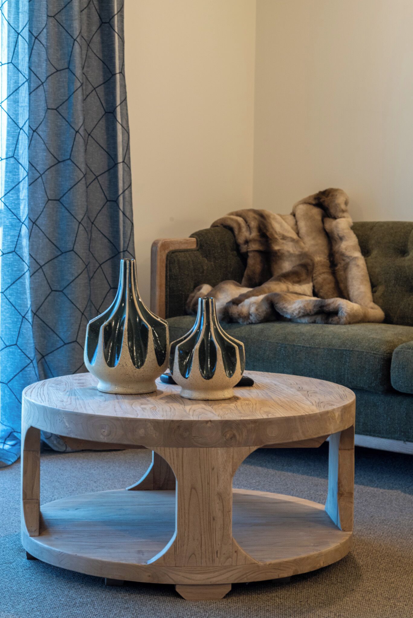 Un tavolo rotondo in legno, due vasi, una tenda grigia con motivo decorativo e un divano verde con una coperta in pelliccia.