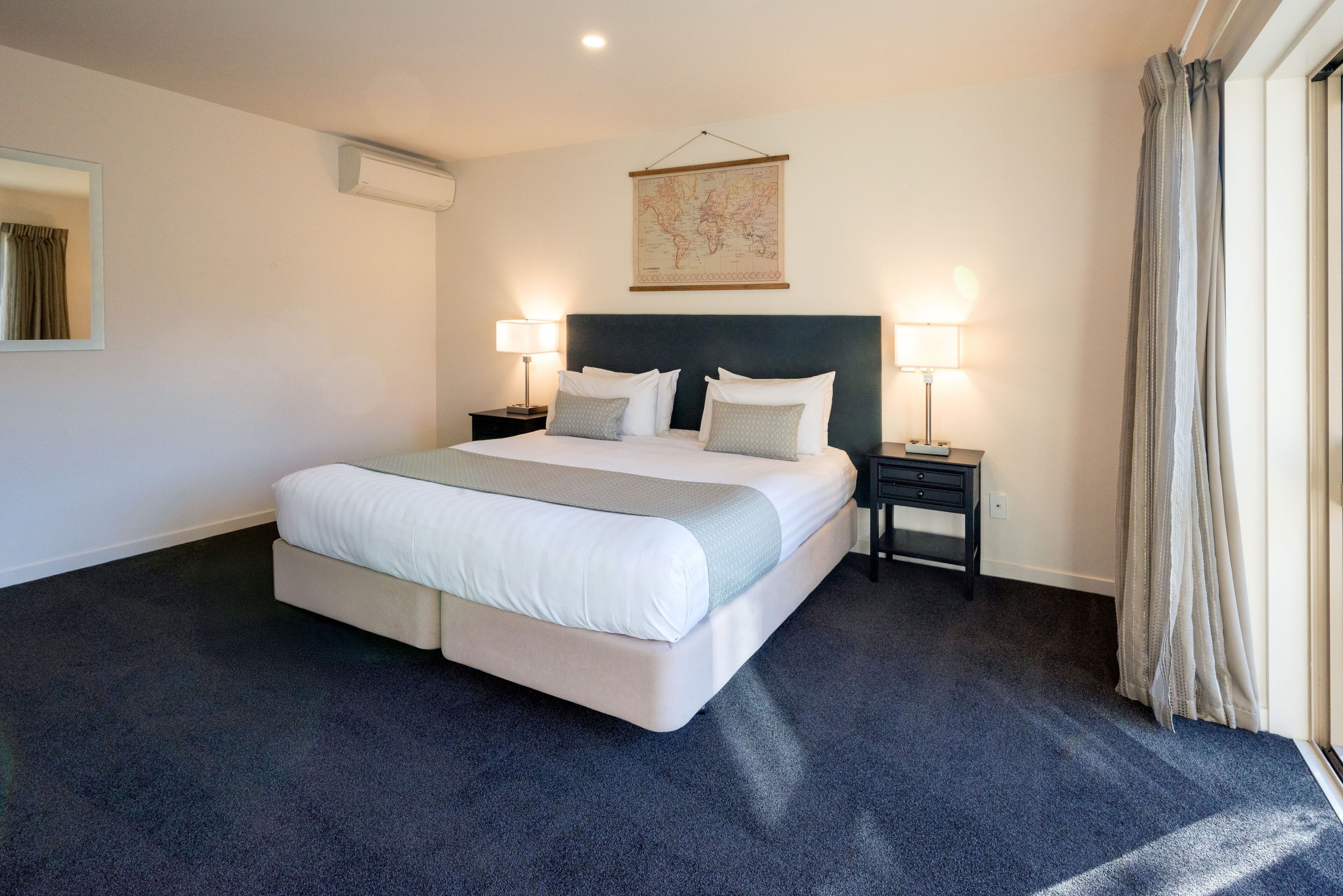 A large bed with a headboard, two bedside tables with lamps, a world map on the wall, and a window with curtains.