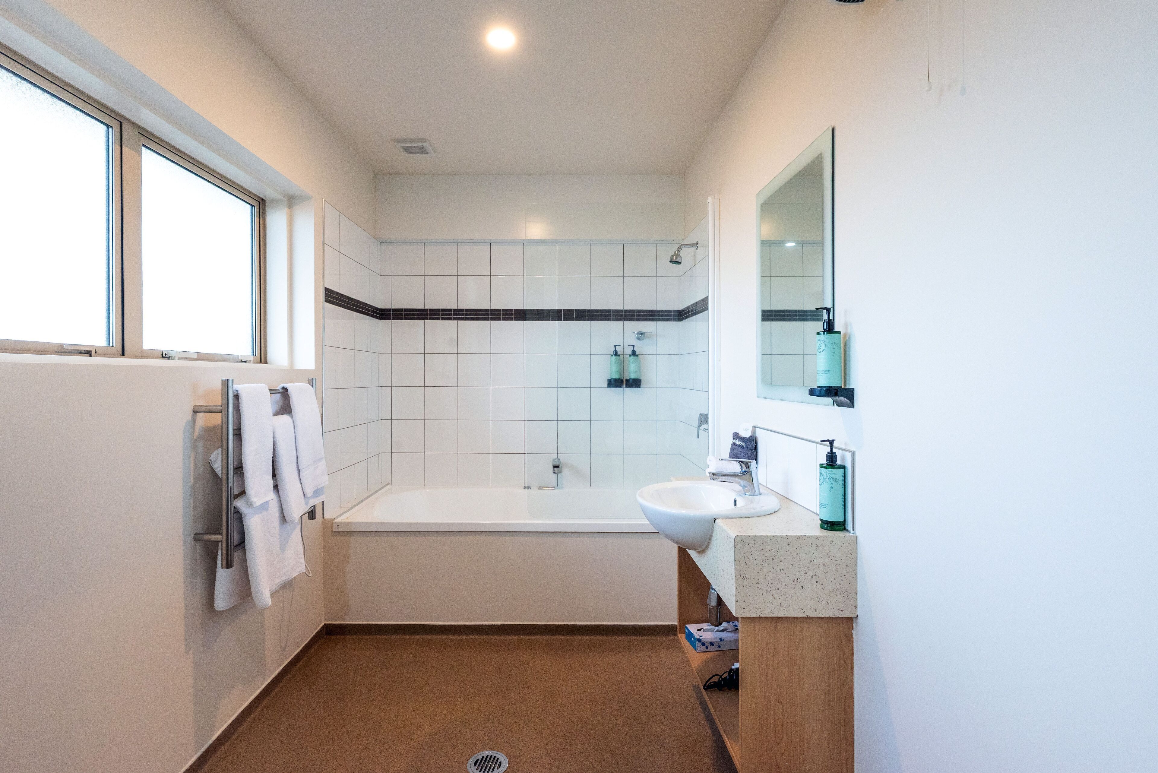 A modern bathroom with a bathtub, a sink, and a large mirror. There are towels hanging on a rack and a window providing natural light.