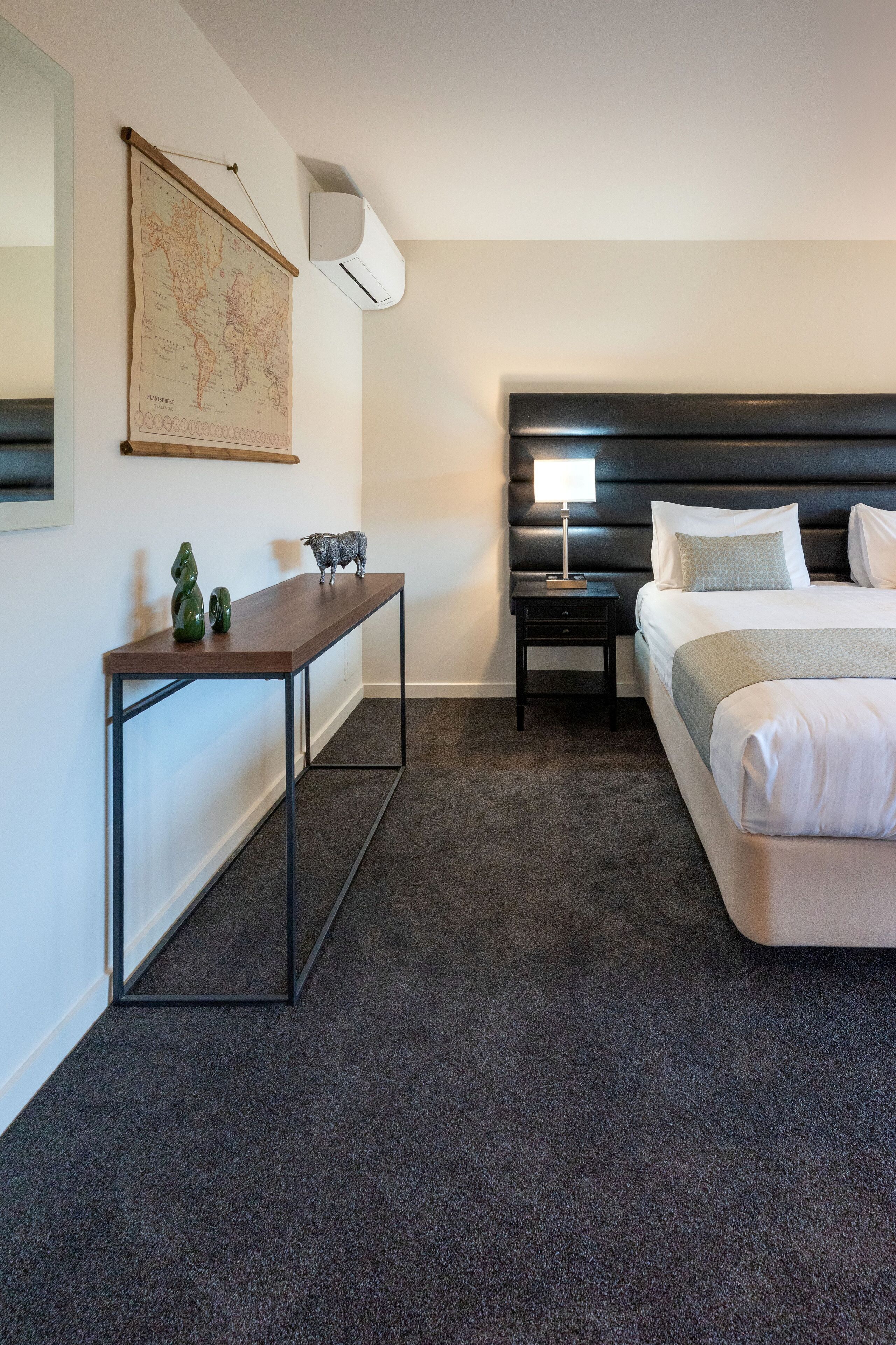 A hotel room with a large bed, a bedside table with a lamp, a small desk with decorative items, and a world map on the wall.