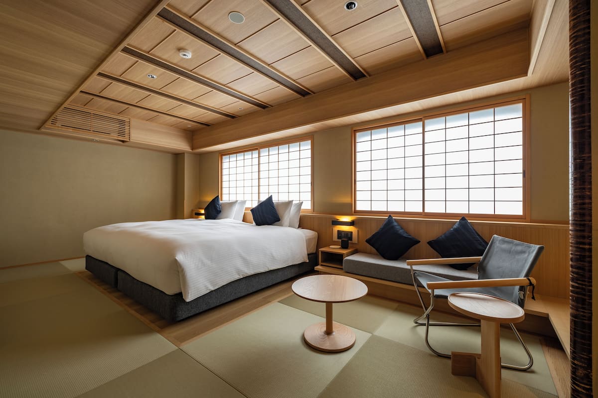 A traditional Japanese-style bedroom with tatami flooring, a bed with a white comforter, a low wooden table, a cushioned bench, and a sofa with cushions.