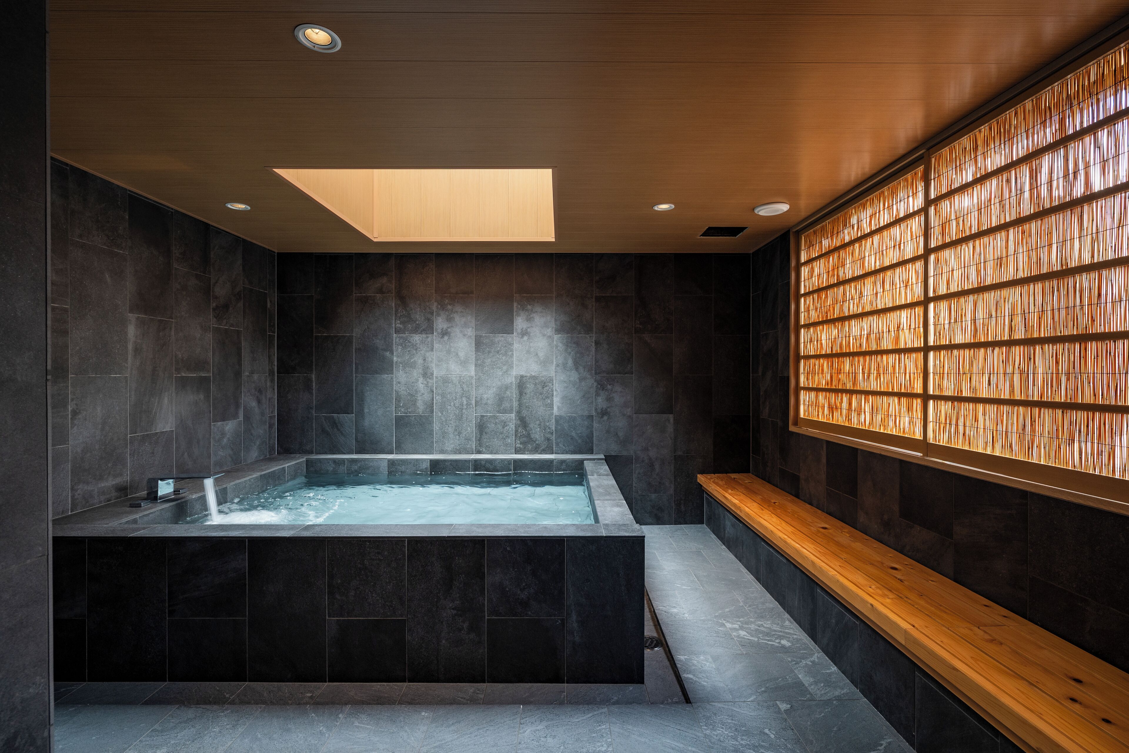 Une salle de spa moderne avec une grande baignoire, des murs en pierre sombre et un banc en bois.