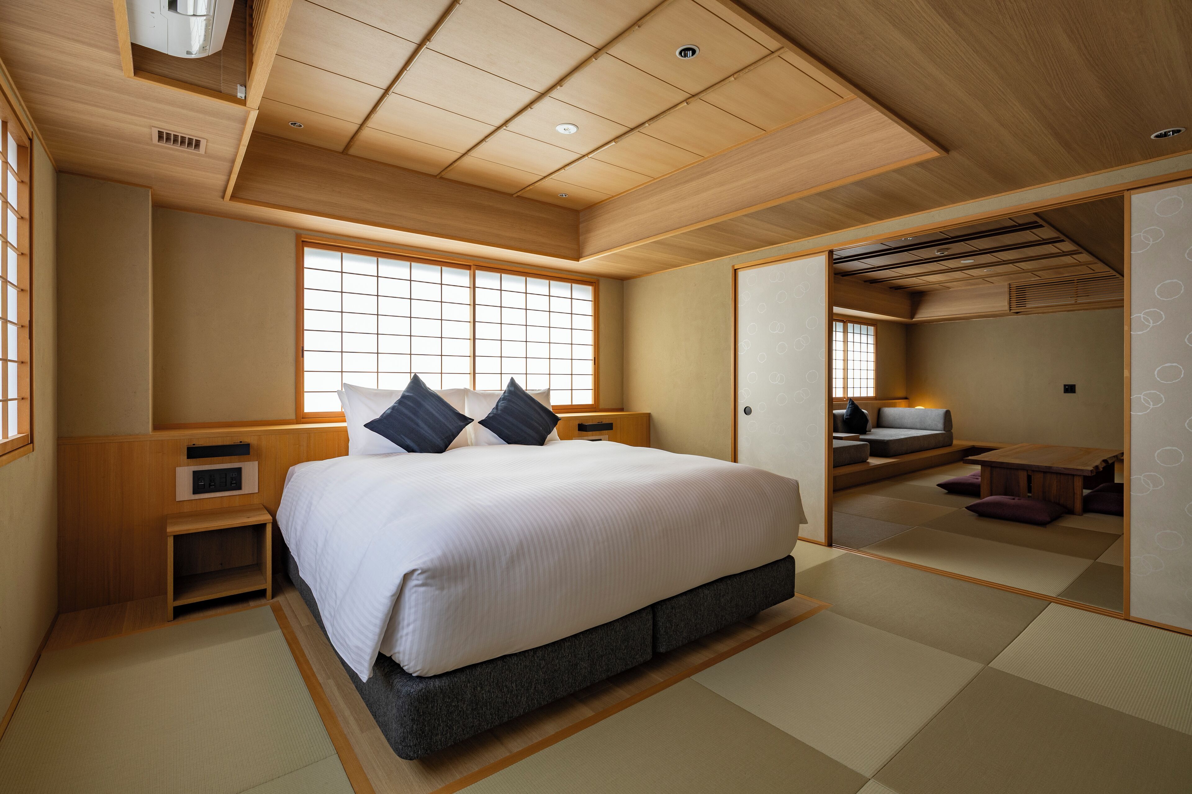 Perinteinen japanilaistyylinen makuuhuone, jossa on tatamimatto, suuri sänky tyynyineen, puinen yöpöytä ja liukuovi, joka johtaa olohuoneeseen, jossa on sohva ja pöytä.