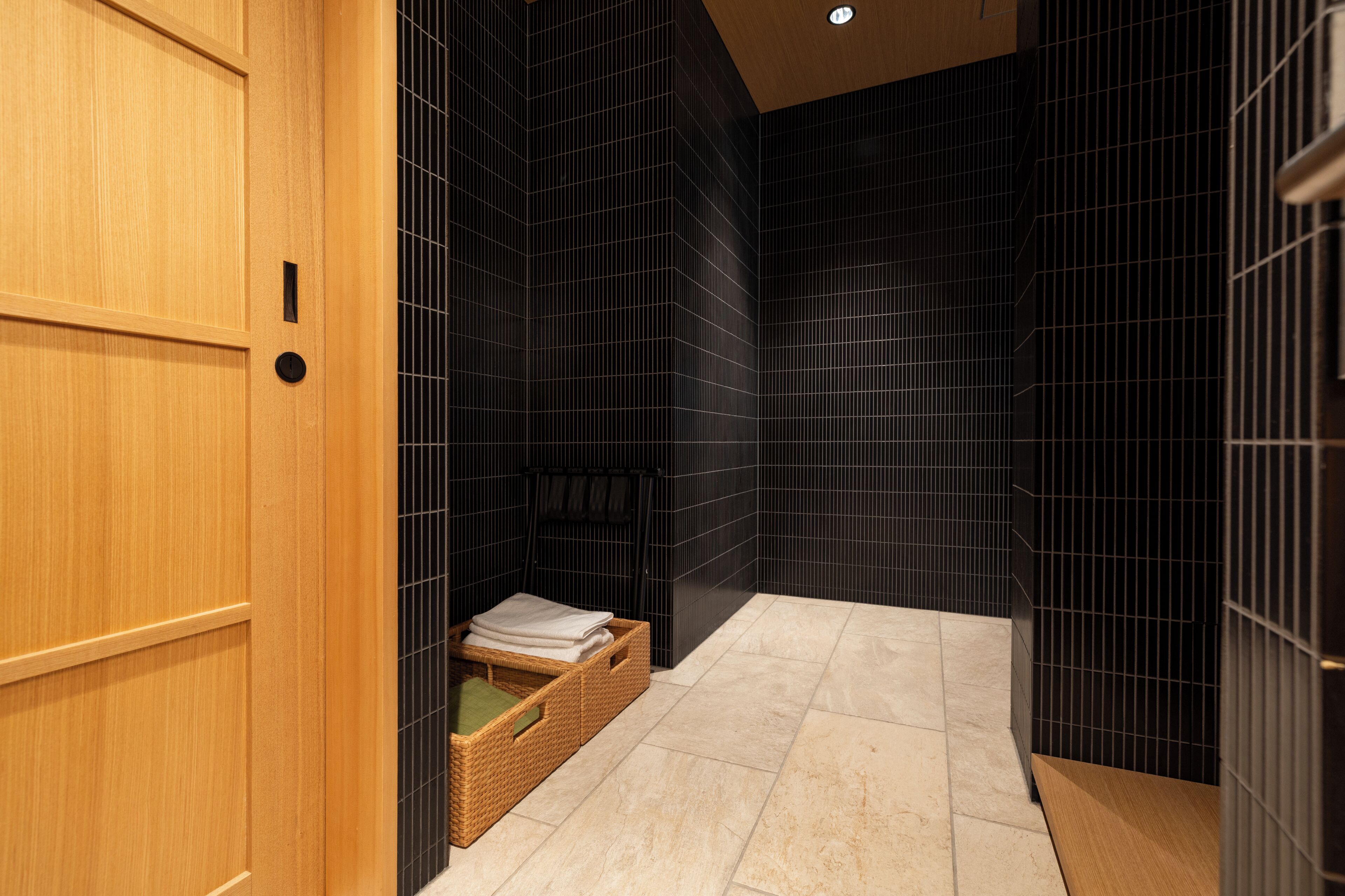 Un baño moderno con paredes de baldosas oscuras, una puerta de madera y una pequeña cesta de mimbre con toallas.