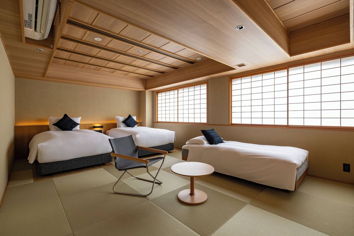 A hotel room with two beds, a small table, a chair, and tatami flooring.