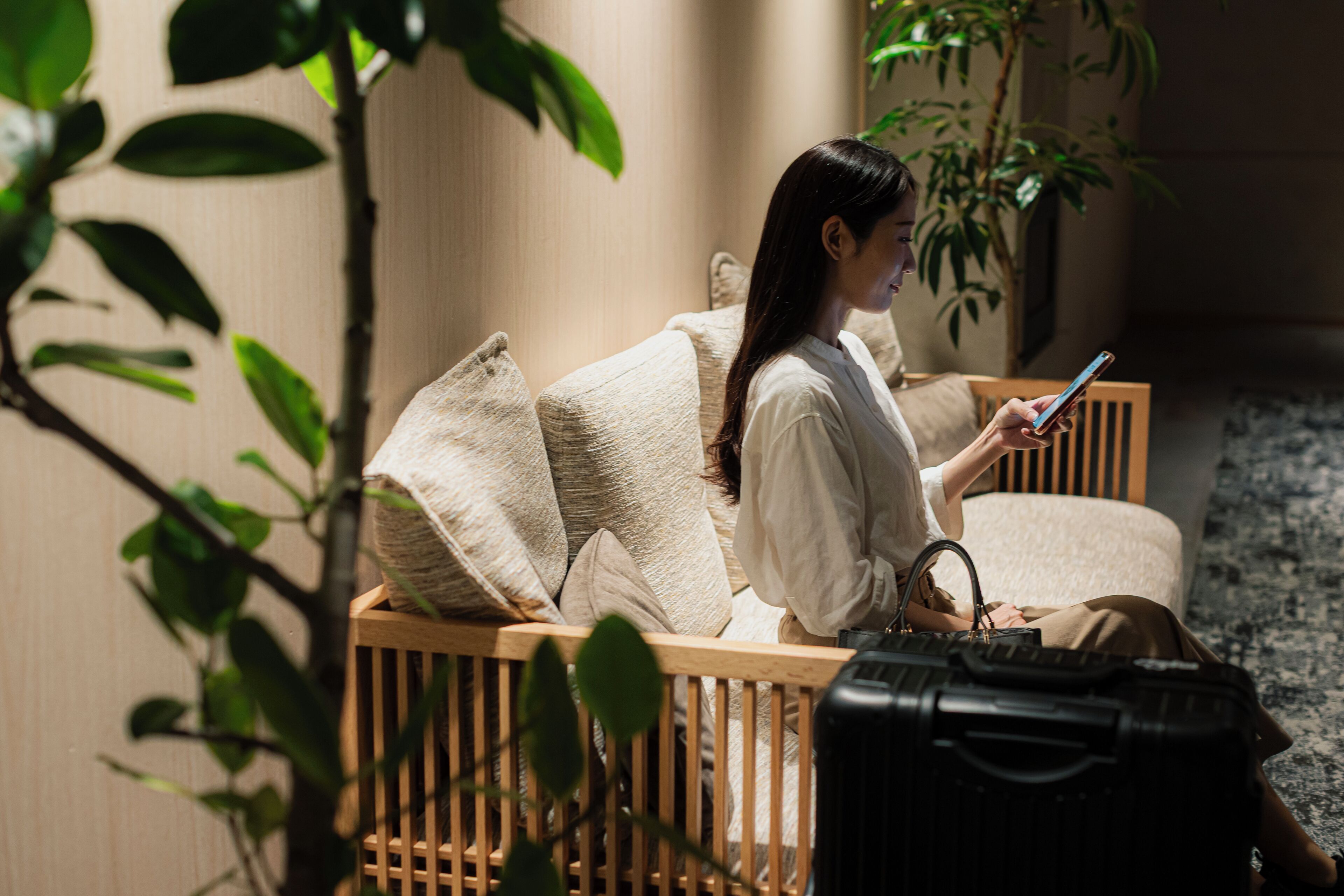A person sitting on a wooden bench with a suitcase, using a smartphone.