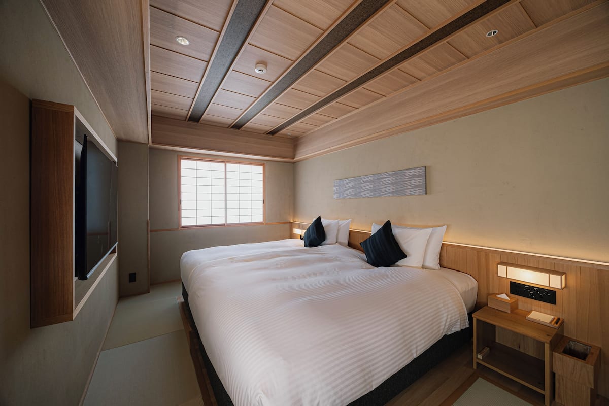 A hotel room with a large bed, wooden headboard, bedside table with lamp, and a TV mounted on the wall.