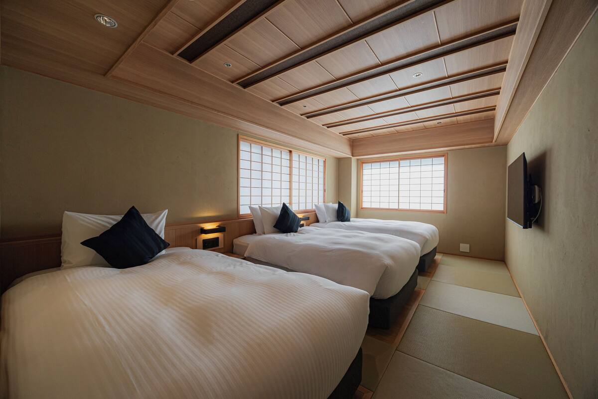 A hotel room with two beds, wooden ceiling beams, large windows, and a flat-screen TV mounted on the wall.