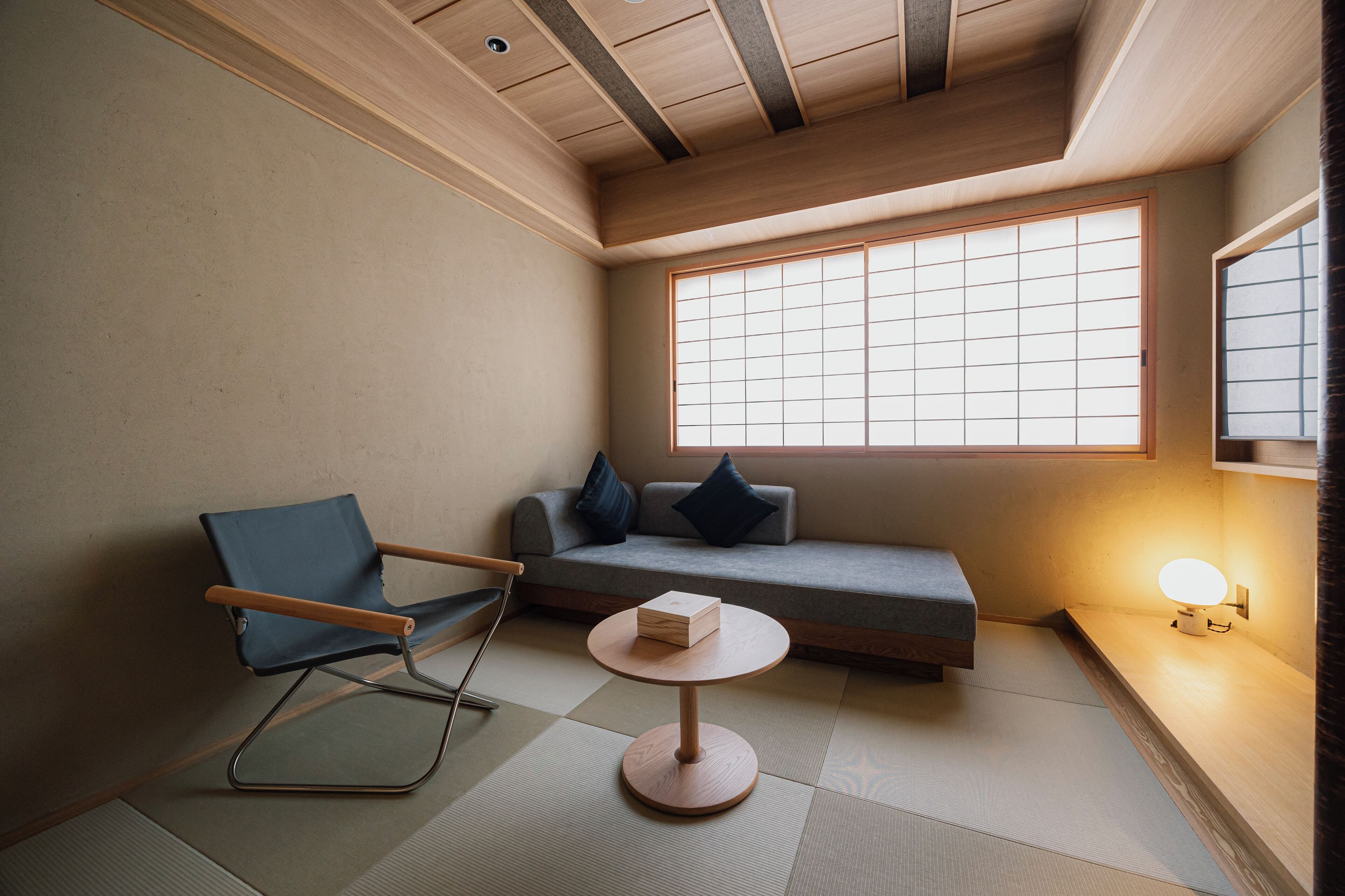 A minimalist room with tatami flooring, a low wooden ceiling, a sofa with cushions, a blue chair, and a small round table with a box on it.