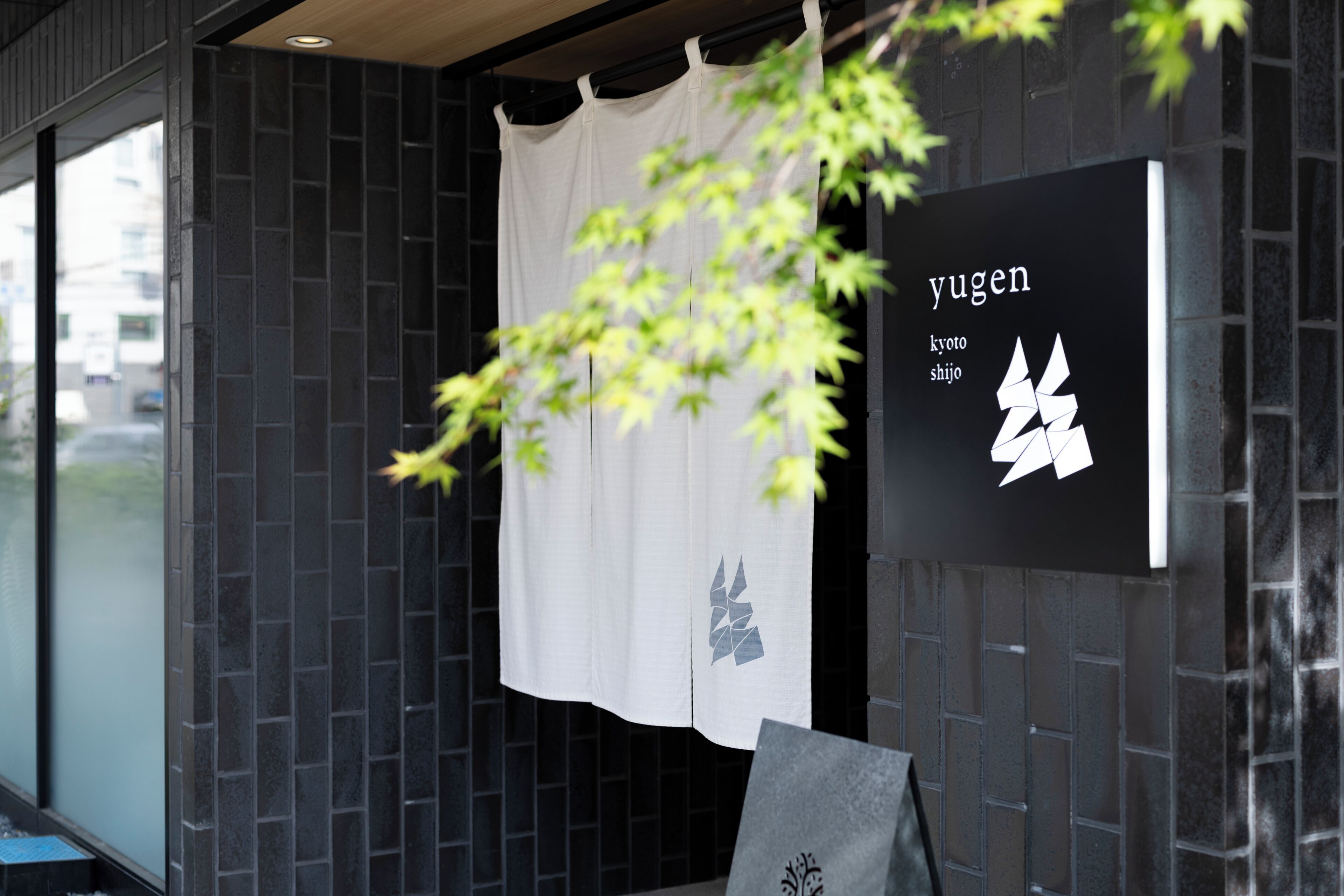 A building facade with a black tiled wall, a white curtain with a geometric design, and a sign with the name 'yugen' and Japanese characters.