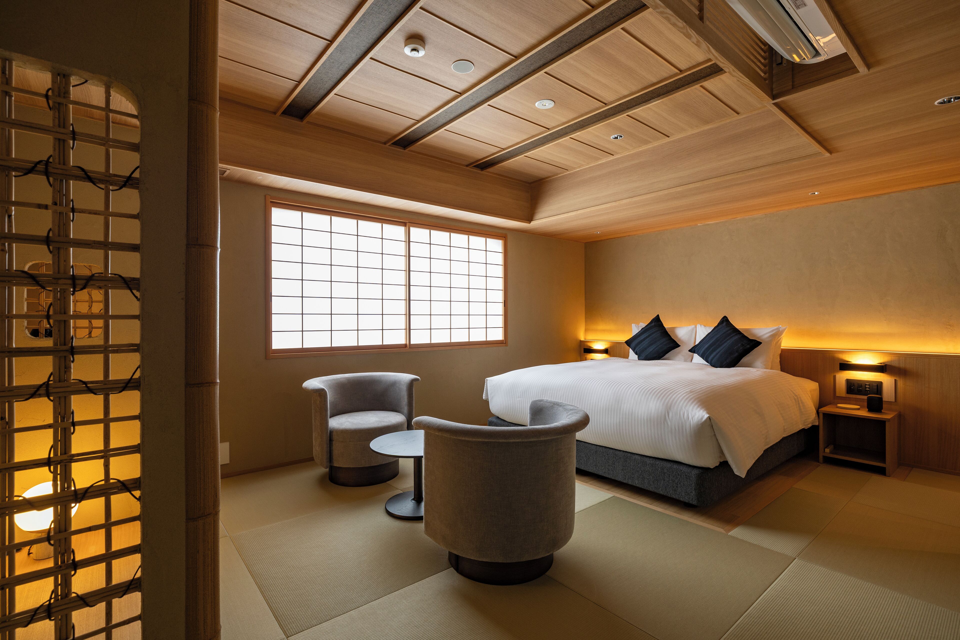 A modern hotel room with a large bed, two armchairs, a small table, and a traditional Japanese sliding door.