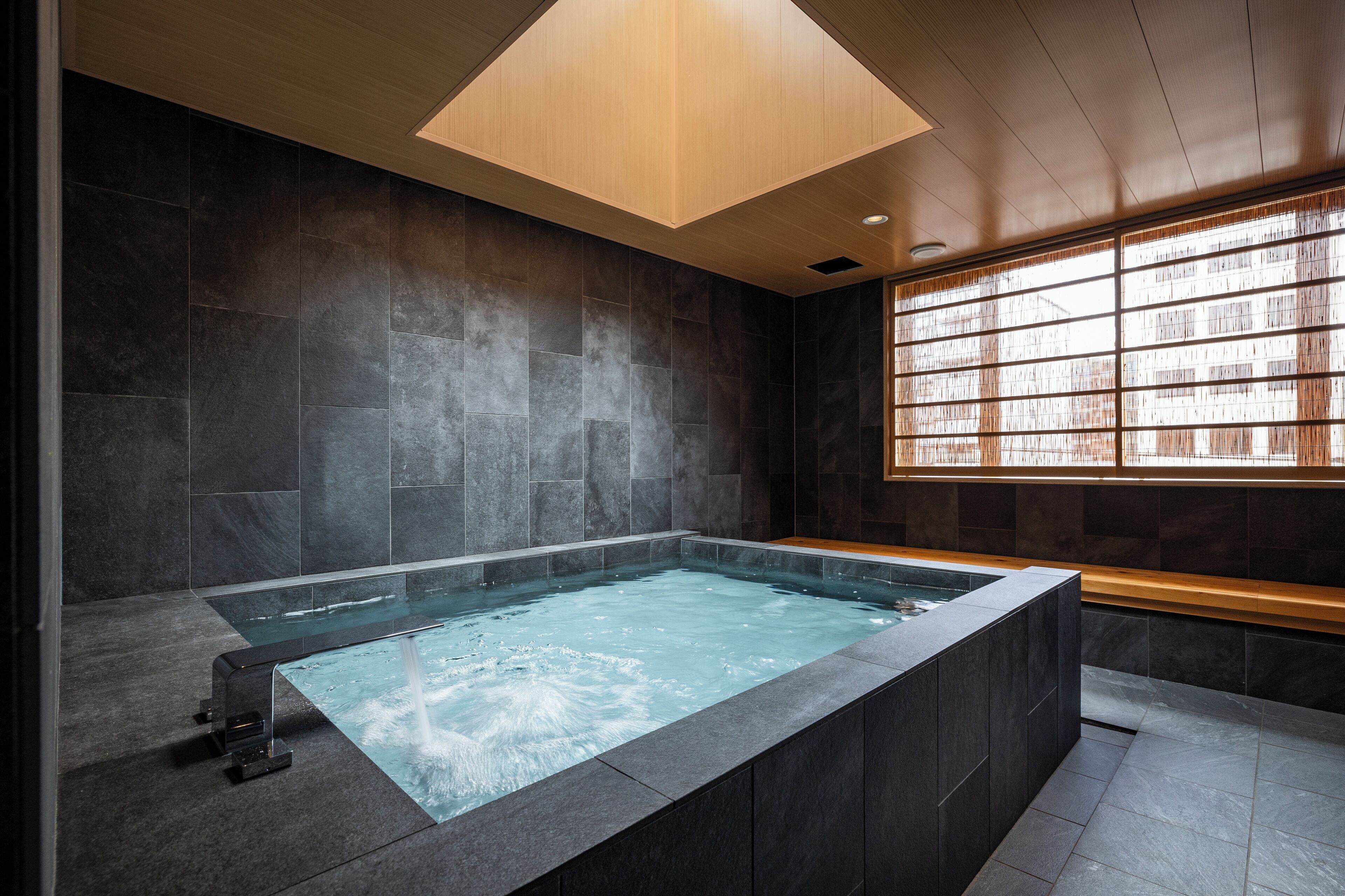 Un moderno cuarto de spa con una bañera de hidromasaje rectangular, paredes de piedra oscura y un banco de madera.