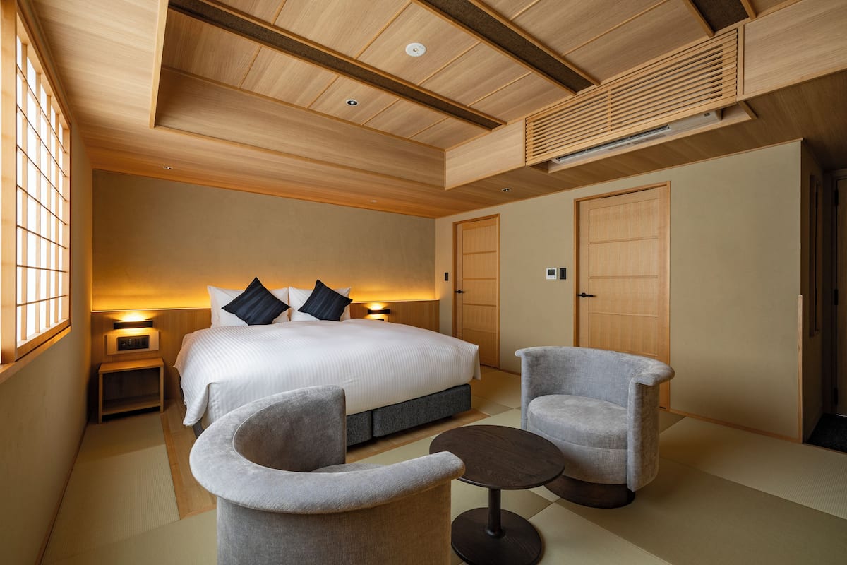 A hotel room with a large bed, two bedside tables, a chair, a small table, and a wooden ceiling.