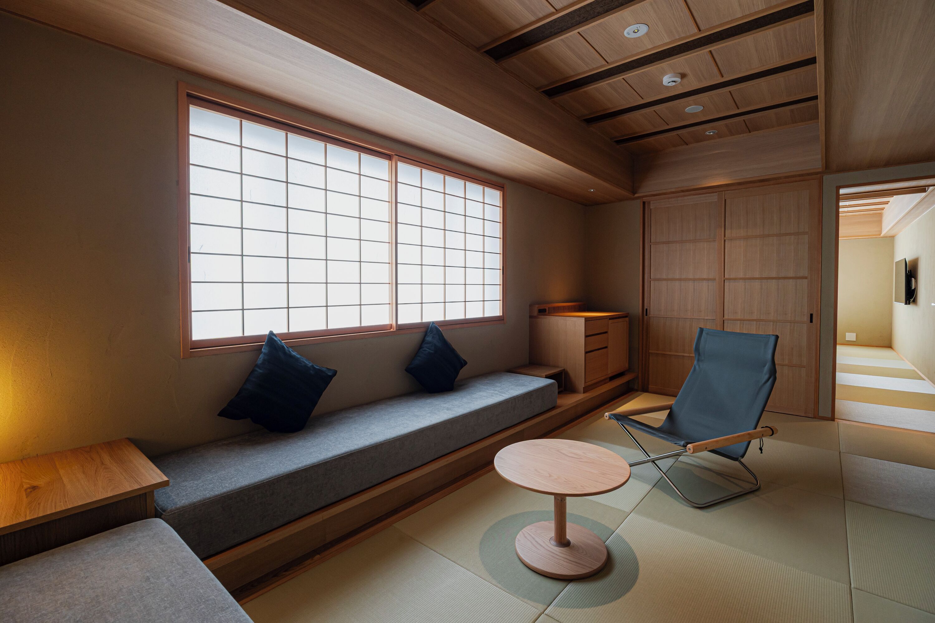 Una habitación japonesa tradicional con suelo de tatami, una mesa de madera baja, un banco acolchado, una silla azul y puertas correderas.
