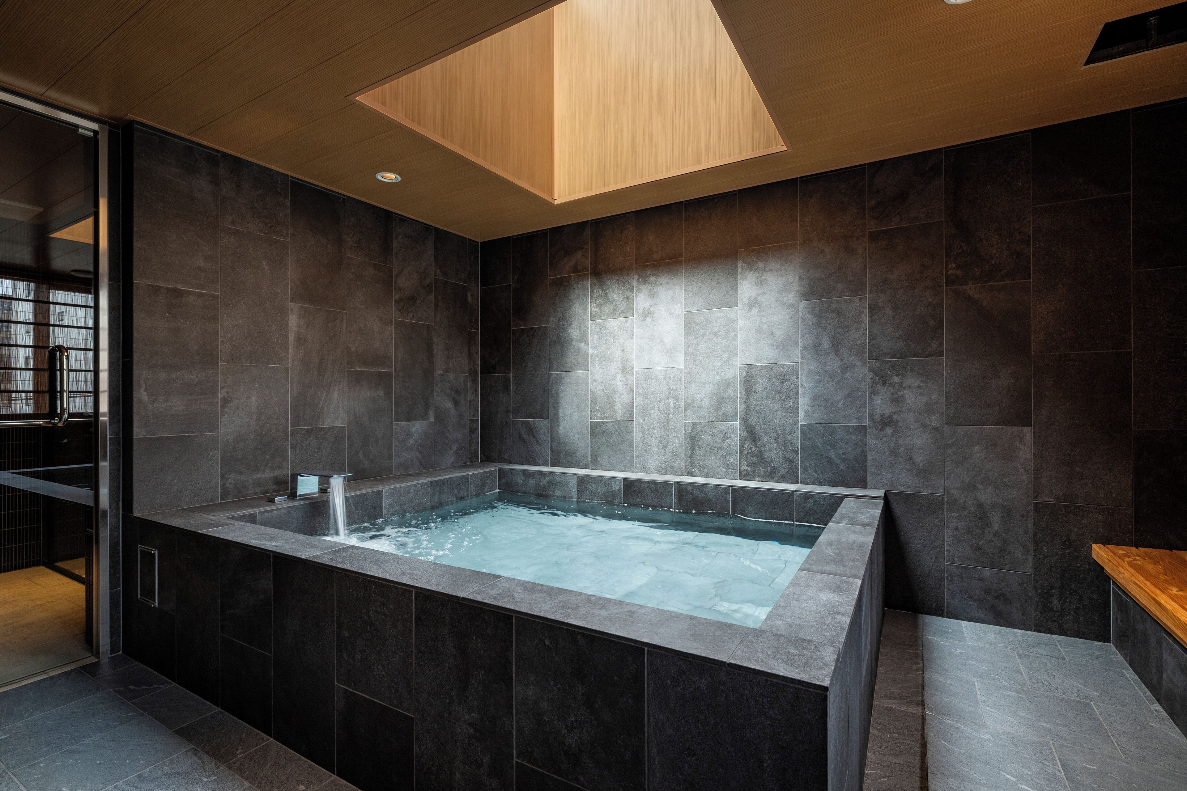 Un baño moderno con una bañera grande, baldosas oscuras y techo de madera.