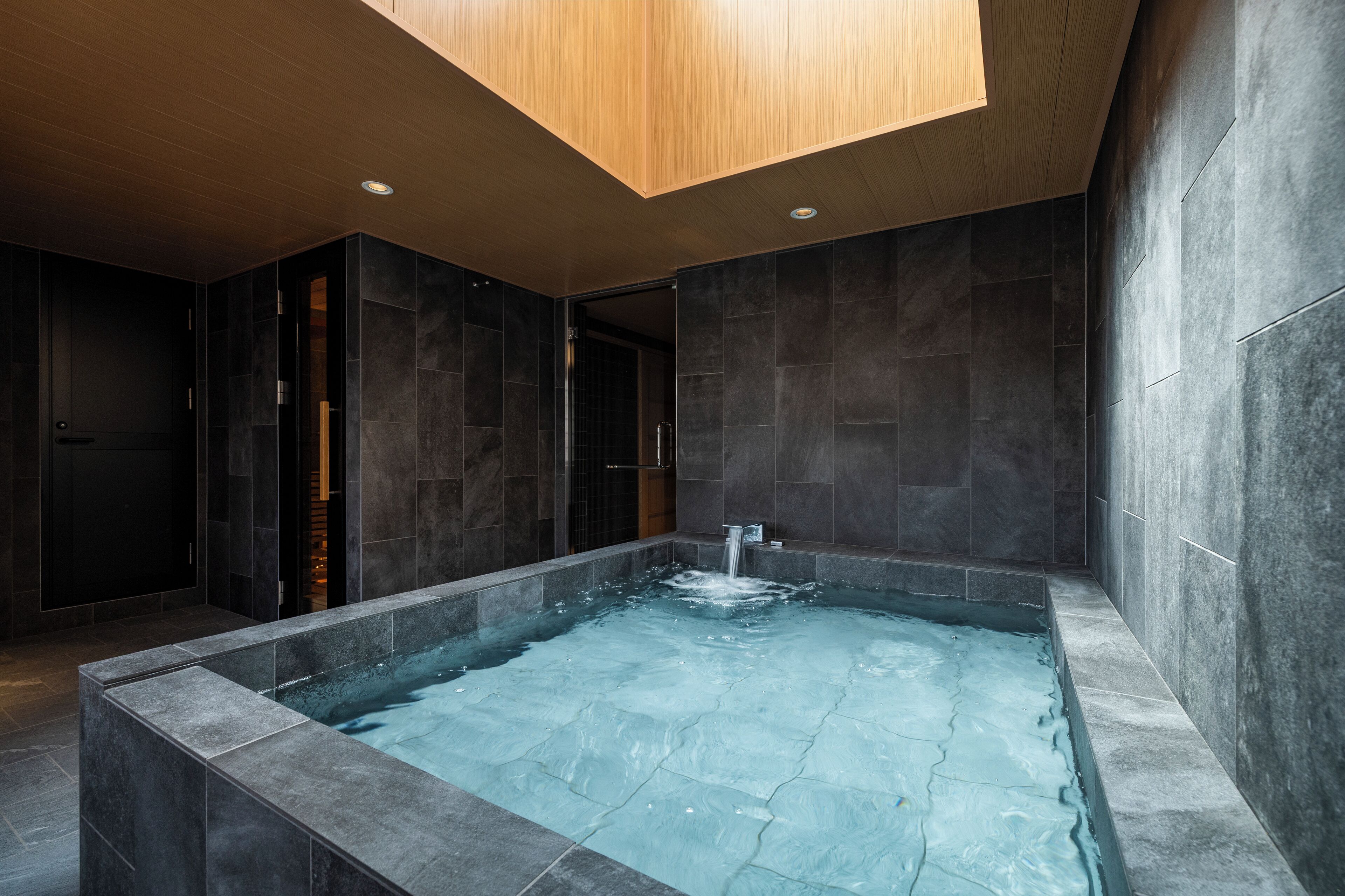 A modern spa area with a rectangular hot tub, dark stone walls, and a wooden ceiling.