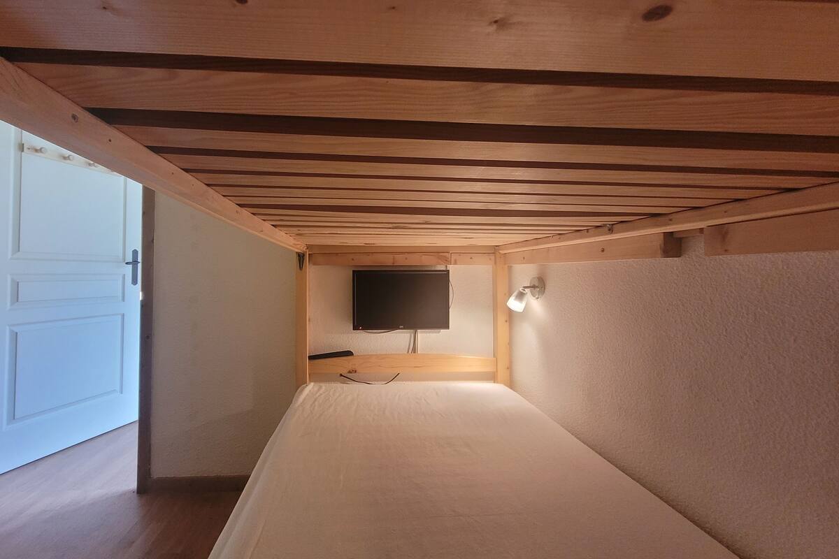 Una habitación estrecha con paneles de madera, una cama, un televisor de pared y una mesita de noche pequeña.