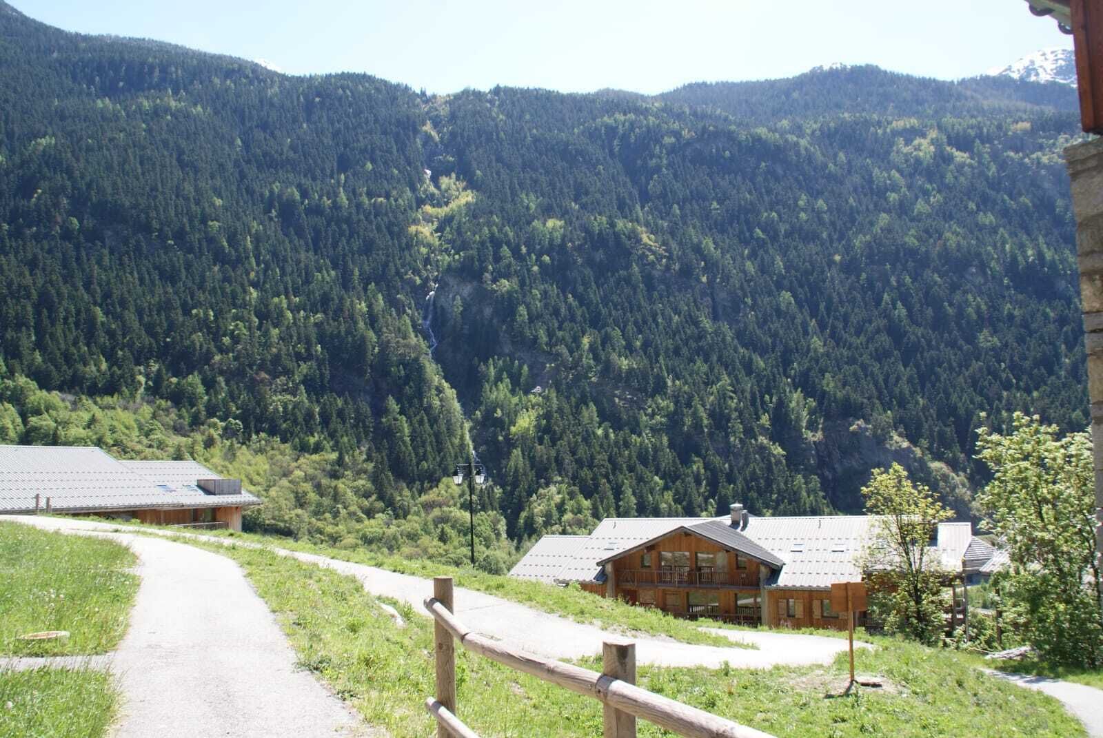 Una cabaña de montaña con un sendero de tierra que conduce a la entrada, rodeada de un bosque denso y colinas.