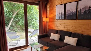 Ein gemütliches Zimmer mit einem dunkelbraunen Sofa, zwei weißen Kissen, einem Glastisch und Blick auf Grün hinter großen Schiebetüren.