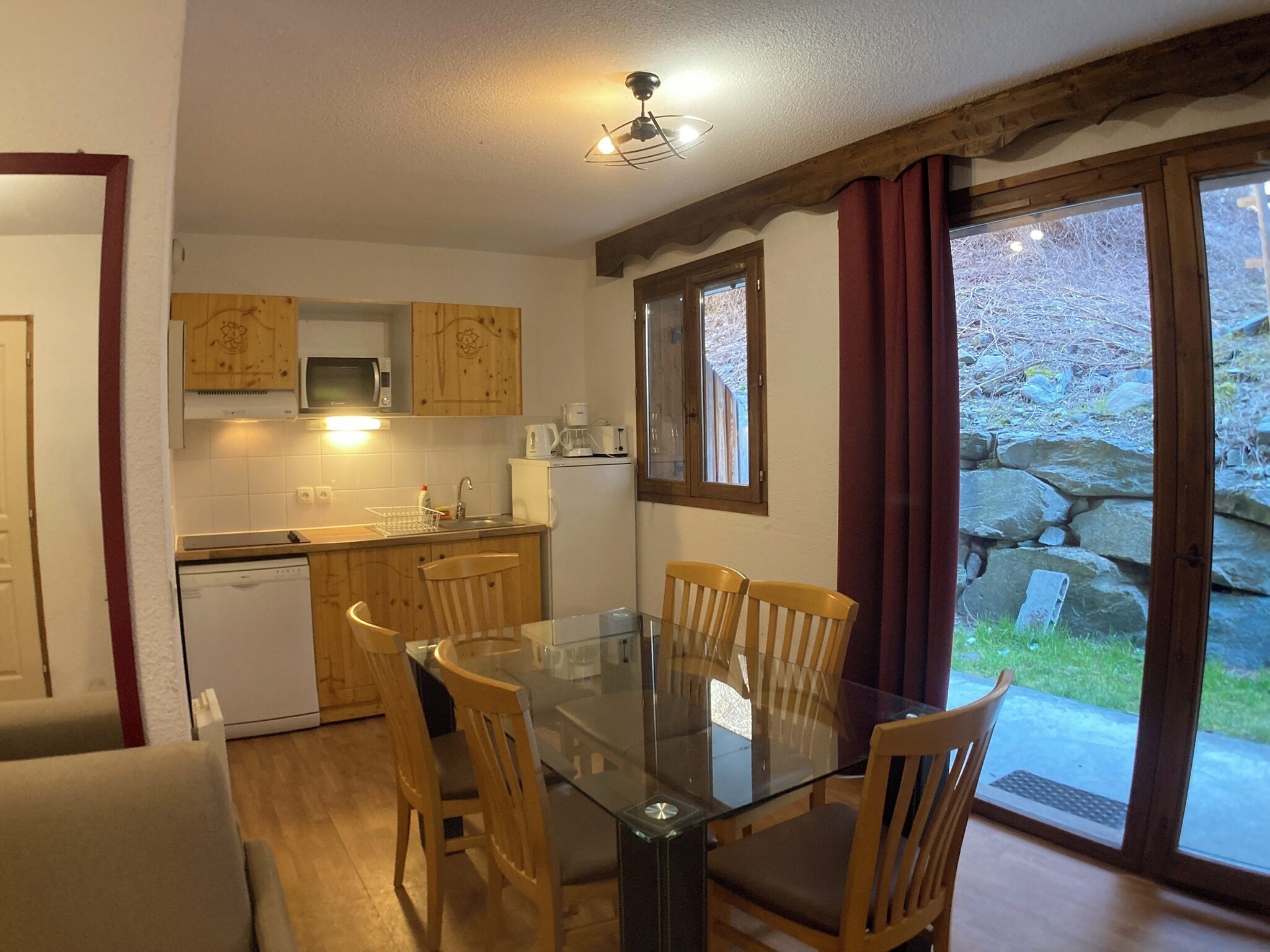 Een compacte keuken met houten kasten, een glazen eettafel en uitzicht op een stenen tuin door een groot raam.