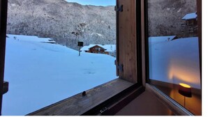 Ein schmales Fenster mit Holzrahmen, durch das man auf eine verschneite Landschaft und ein kleines Blockhaus blickt.