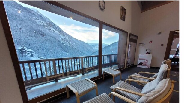 Ein Zimmer mit Balkon und Blick auf einen verschneiten Berg. Mehrere gepolsterte Stühle und kleine Tische sind vorhanden.
