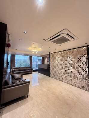 A modern hotel lobby with a reception desk, a sofa, a decorative partition, and a large window offering a city view. - Hotel Presidency (Surat)