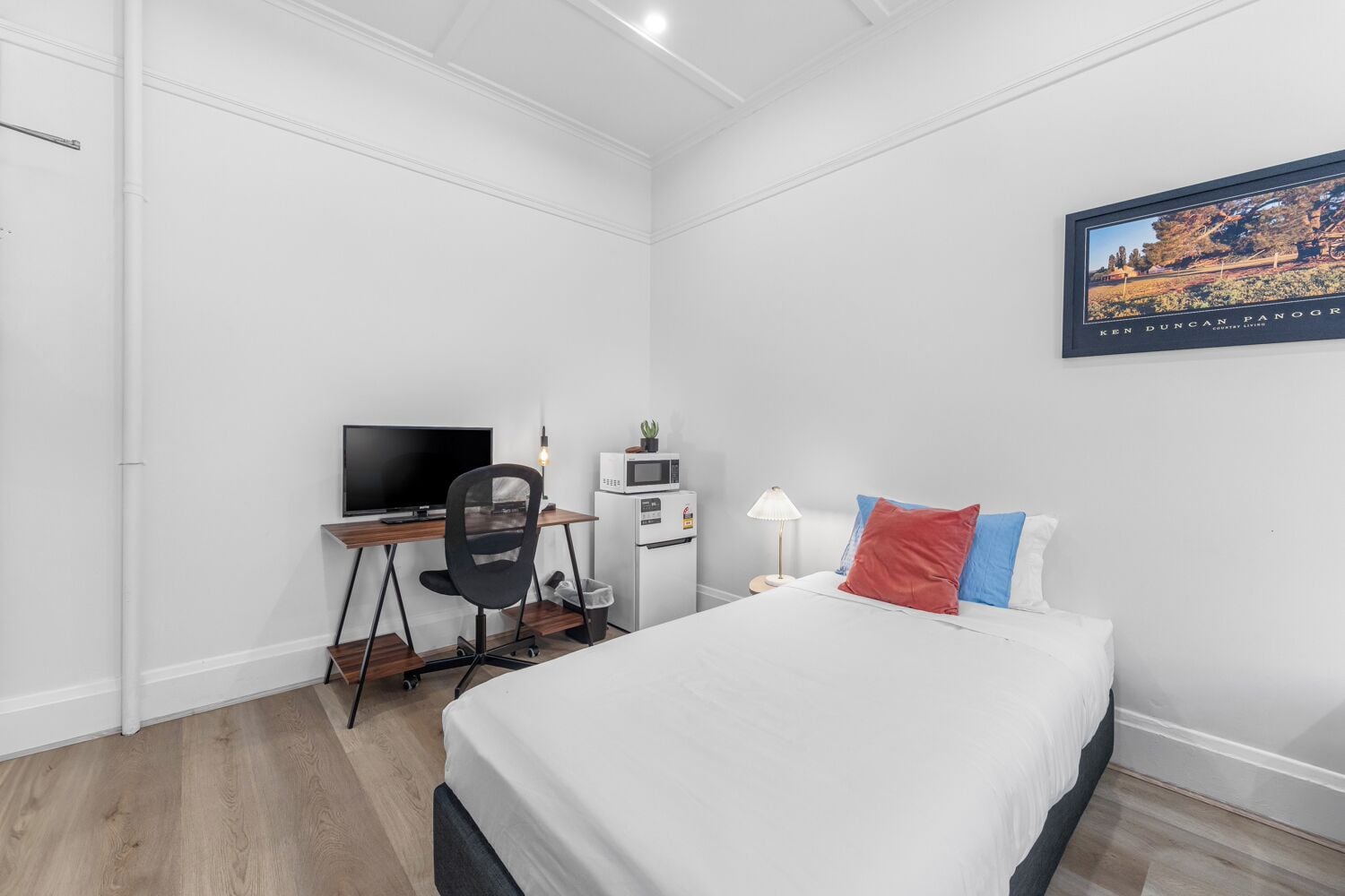 A hotel room with a bed, a desk with a computer, a TV, a microwave, and a small plant.