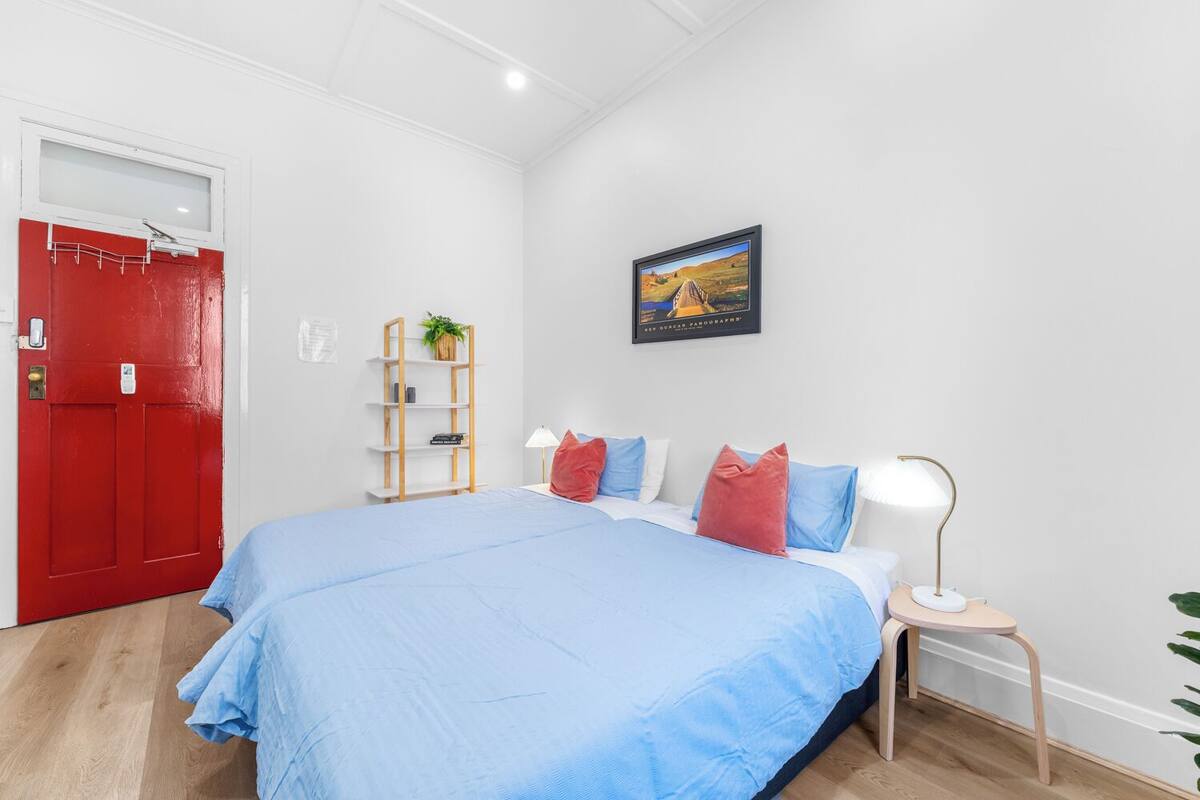 A bedroom with a large bed, a red door, a white wall, a small table with a lamp, and a framed picture on the wall.