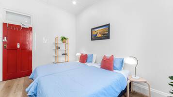 A bedroom with a large bed, a red door, a white wall, a small table with a lamp, and a framed picture on the wall.