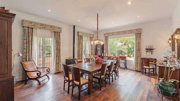 A spacious dining area with a large wooden table and chairs, surrounded by windows with floral curtains.
