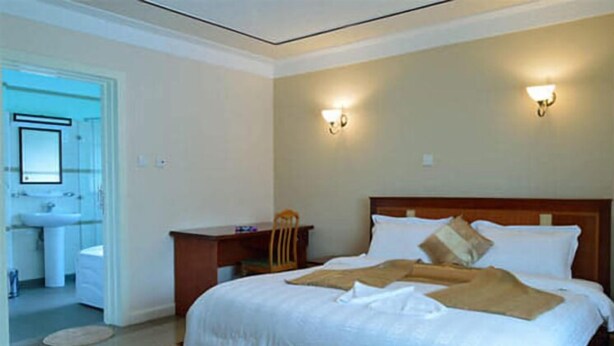 A hotel room with a large bed, a wooden desk, a chair, a nightstand, and a bathroom visible through an open door. - Westlands Gardens Apartment (Nairobi)